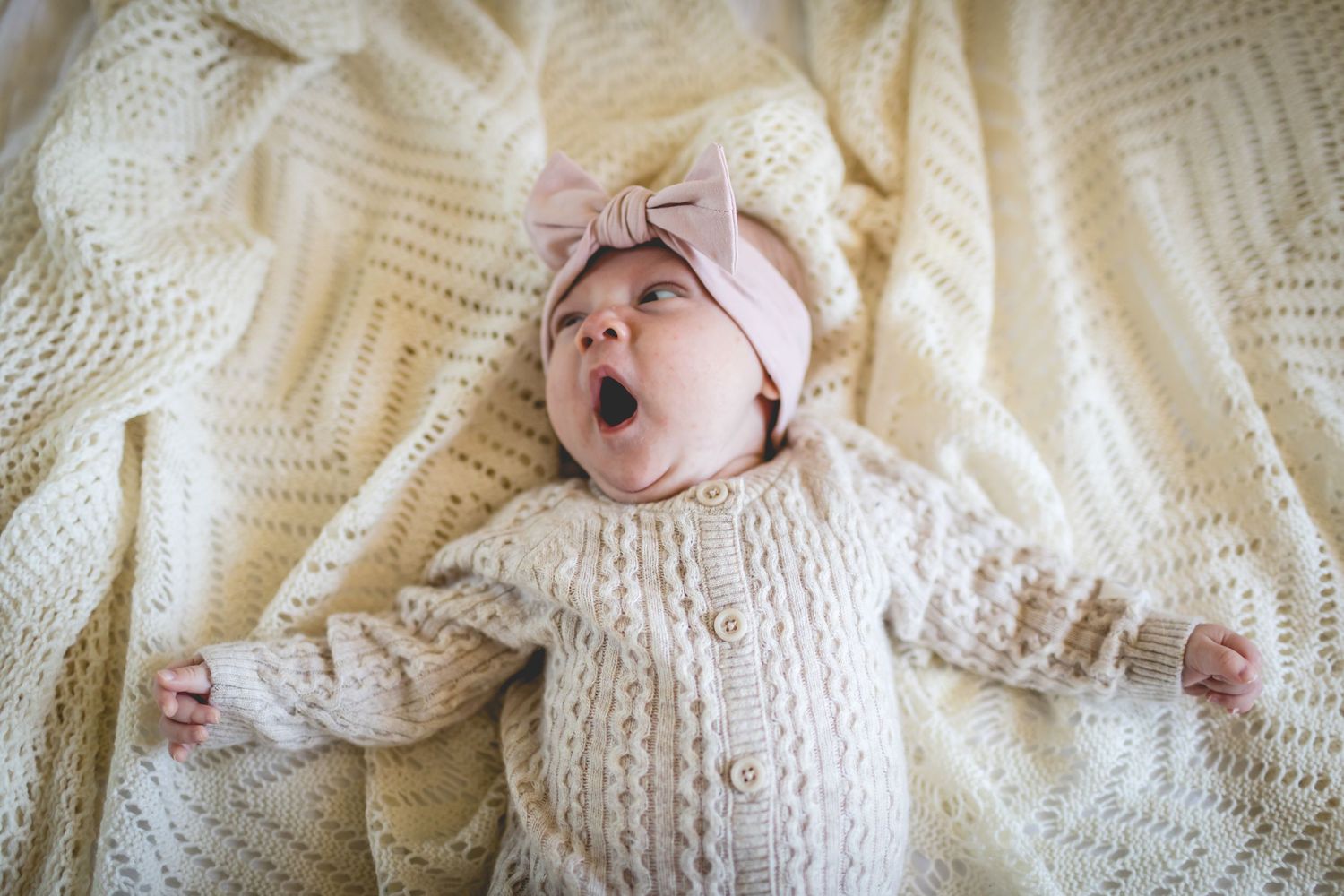 Infant in white knit sweater lying on cream blanket with expressive yawning gesture.