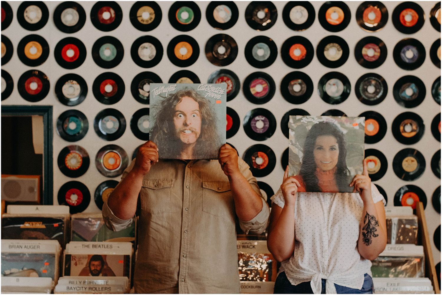 Two people holding vinyl records in front of their faces against a wall decorated with vintage record albums.