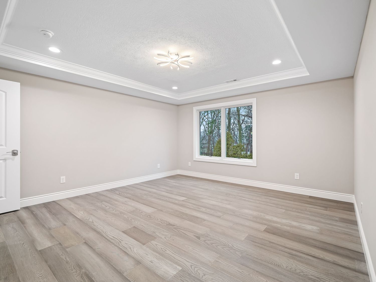 Spacious empty bedroom with tray ceiling, wood flooring, and window overlooking trees.