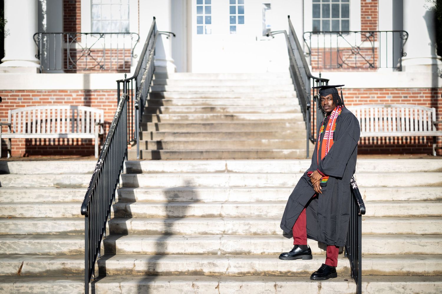 Graduate in red and black regalia posing on grand stone steps of academic building.