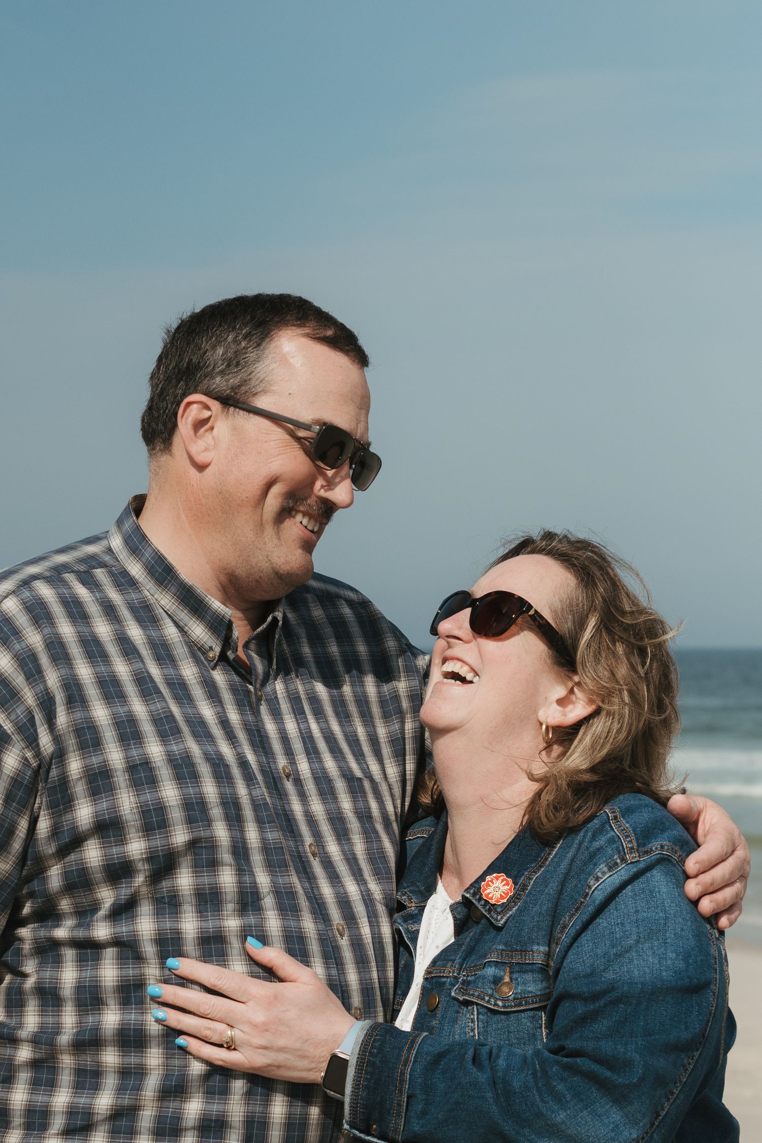 A couple shares a candid laughing moment together by the ocean wearing casual clothes.