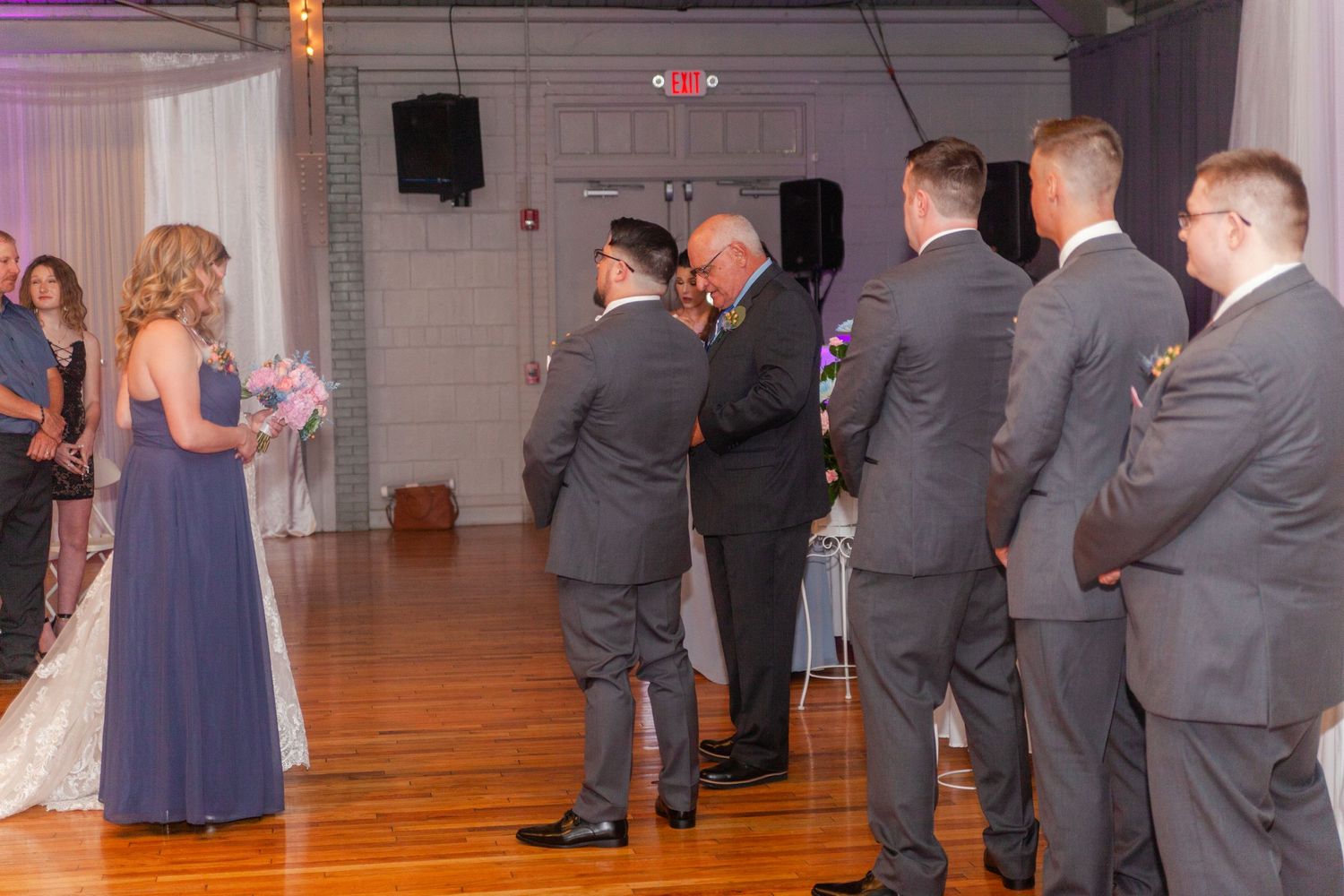 Wedding party in gray suits watching a special moment on the dance floor at an indoor reception venue.