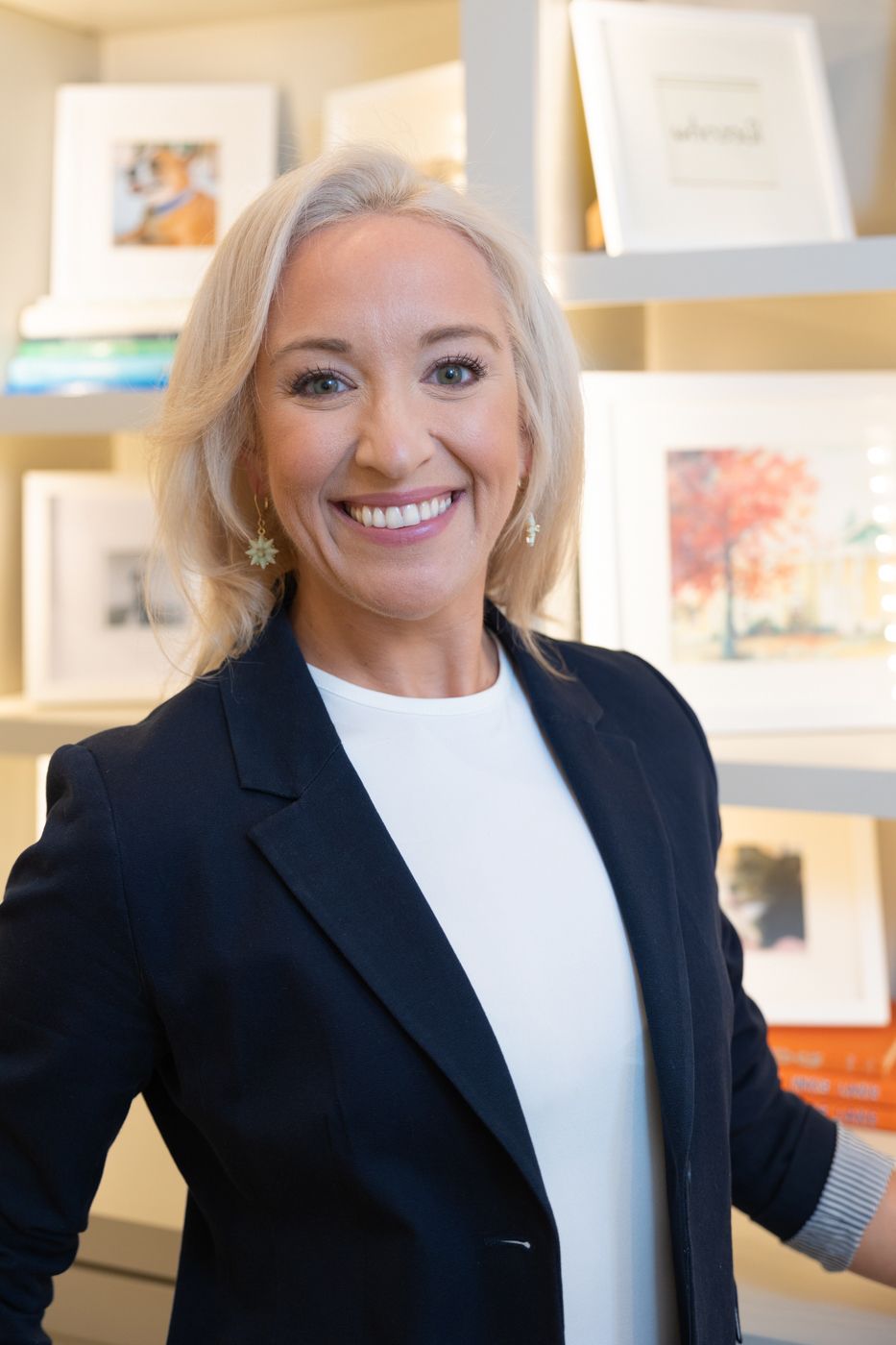 Professional woman in black blazer and white top poses against backdrop of framed artwork.