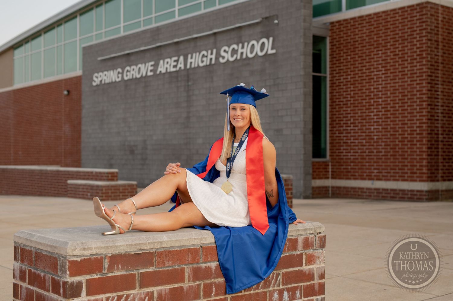 Graduate in blue and red regalia sits on brick ledge outside Spring Grove Area High School.