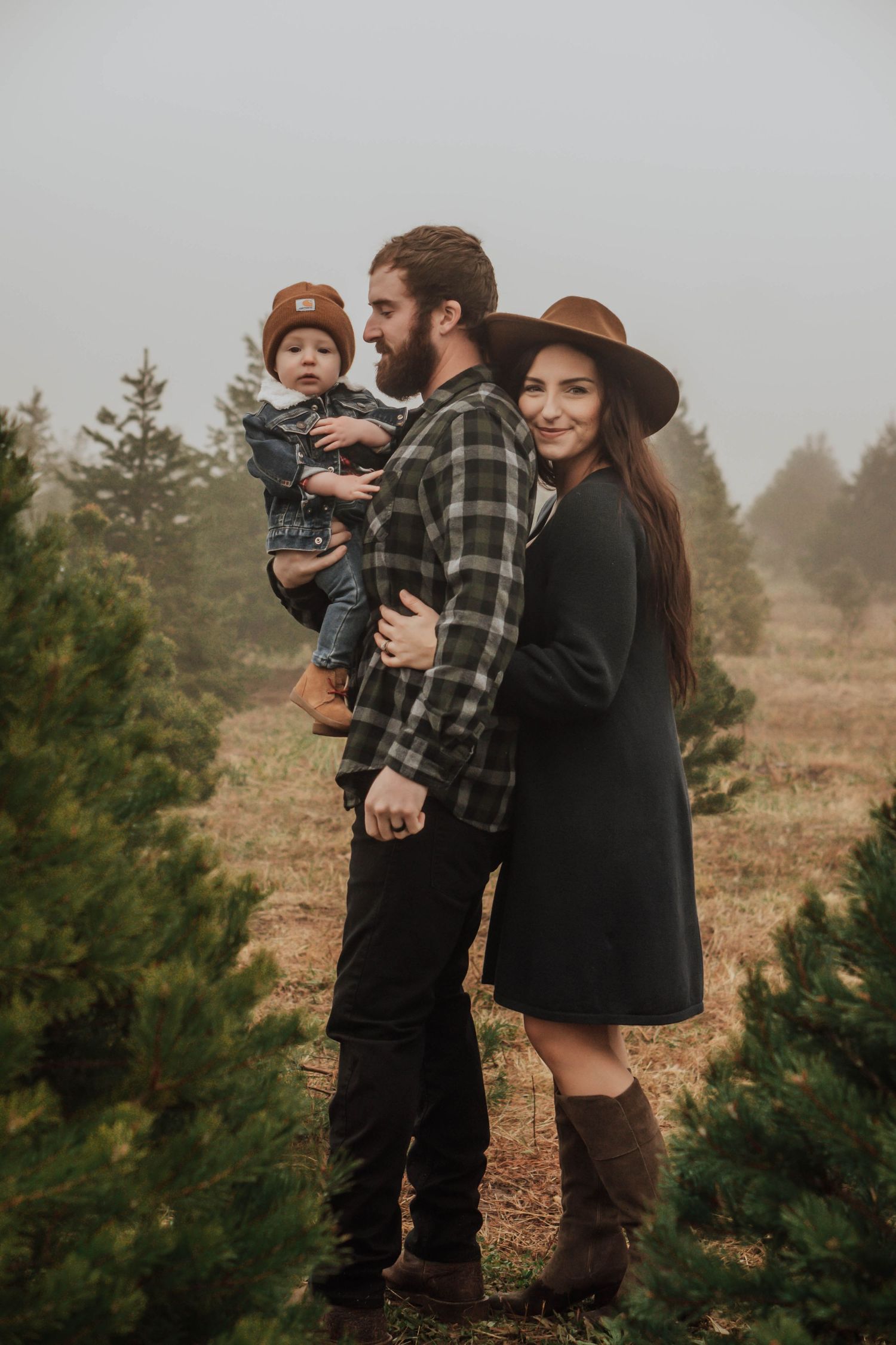 Young family with baby at misty Christmas tree farm in fall attire.