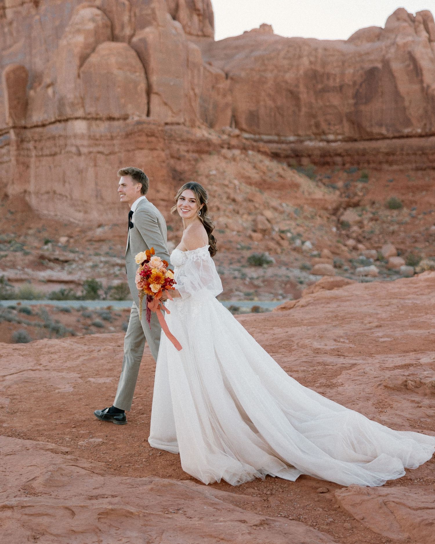 High-end editorial destination wedding photography featuring a bride and groom walking in a majestic red rock desert.