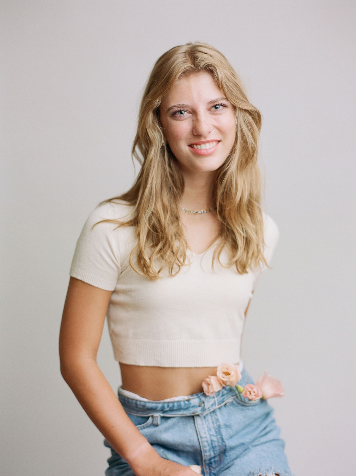 teenage girl in ivory crop top sweater and denim cut offs with flowers in pockets against neutral backdrop