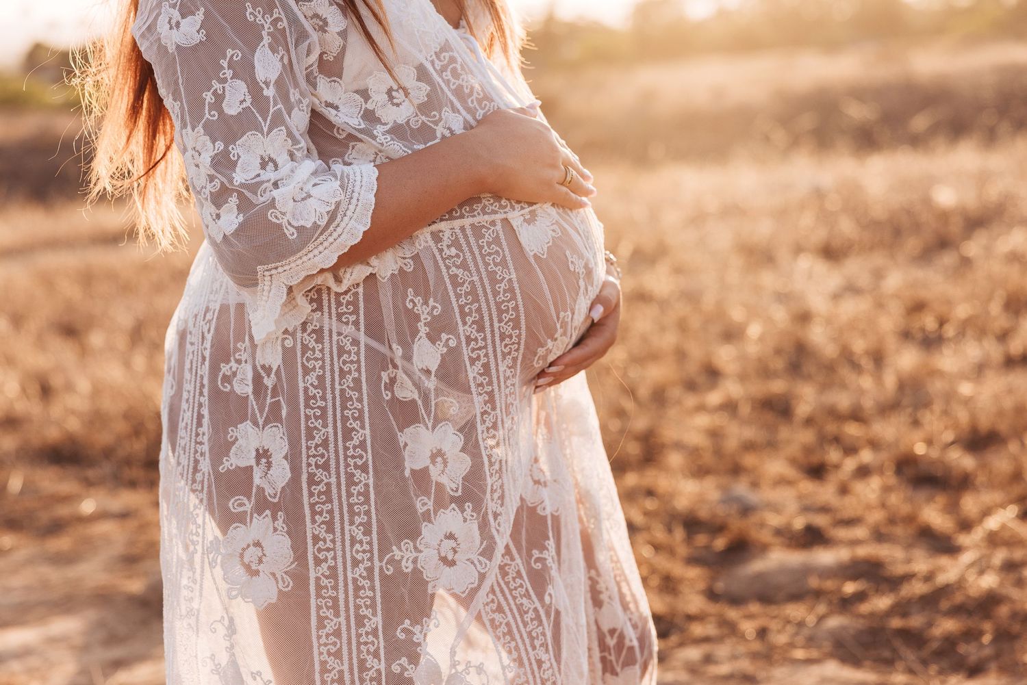 Soft focus shot of pregnant belly wrapped in intricate white lace dress against golden sunset.