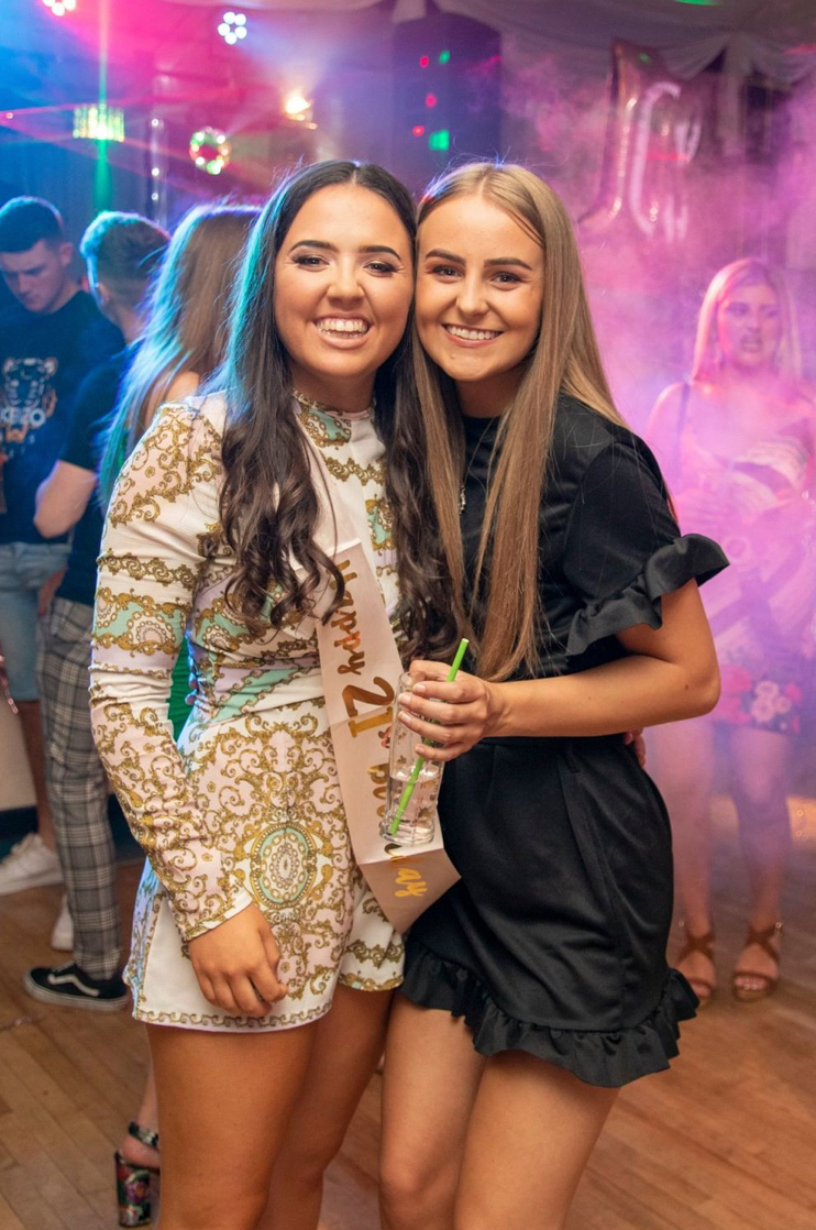 Two young women smiling and posing together at a colorful birthday party celebration.