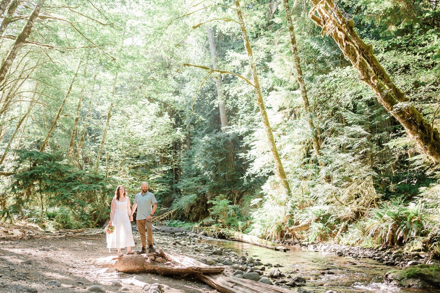 A couple walks next to a creek through lush green trees with dappled sunlight filtering through the canopy.