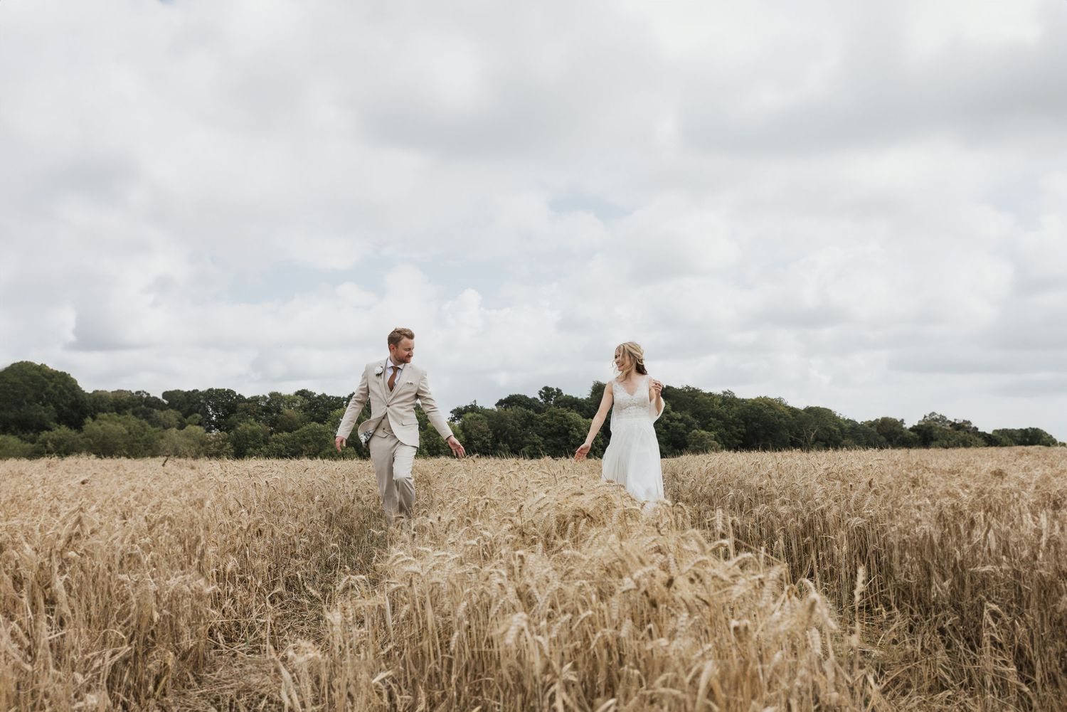 Rob and Abbie are walking through a wheat field and reach out their hands to each other wearing a suit and a white dress