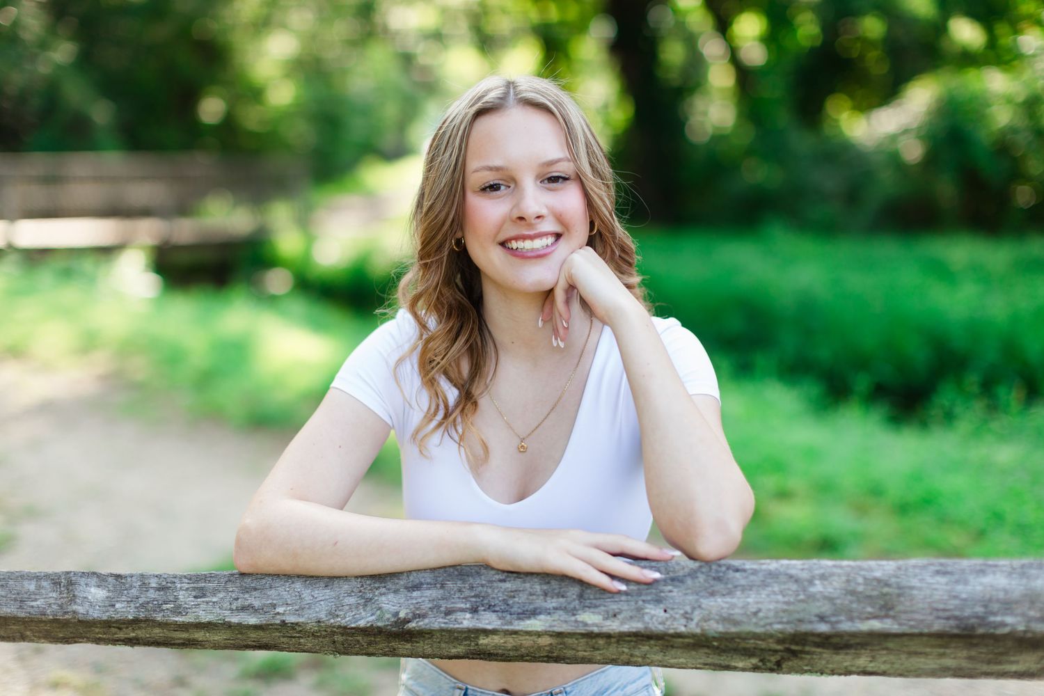 Someone in a white top poses casually by a wooden fence on a sunny day with lush green foliage in the background.