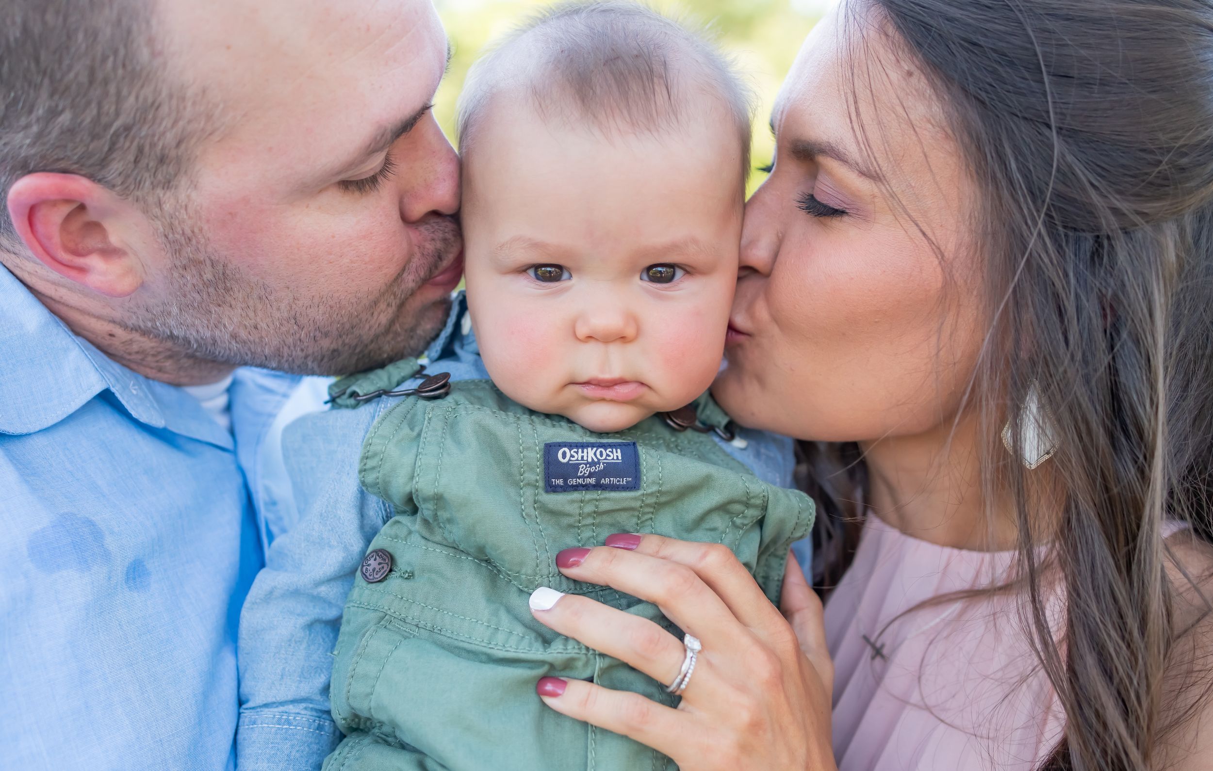 Sweet family moment as parents kiss baby in green outfit against soft background.