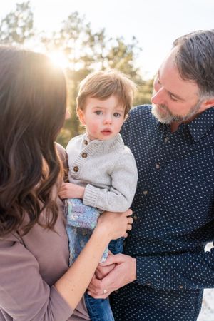 Intimate family portrait captured outdoors with soft sunlight streaming through trees in the background.