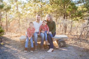 Family of three sitting together on a rustic bench surrounded by pine trees in autumn.