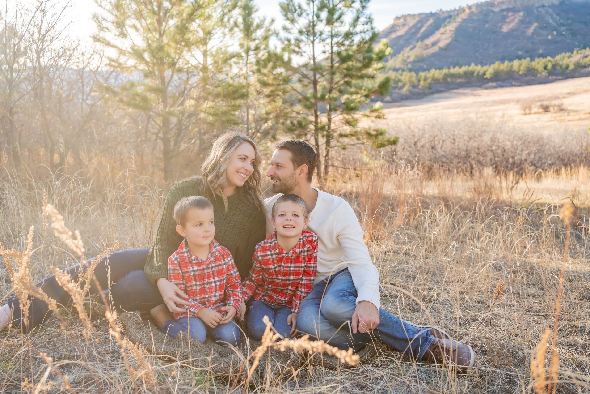 Family sitting together in golden grass field with mountains and pine trees in background.