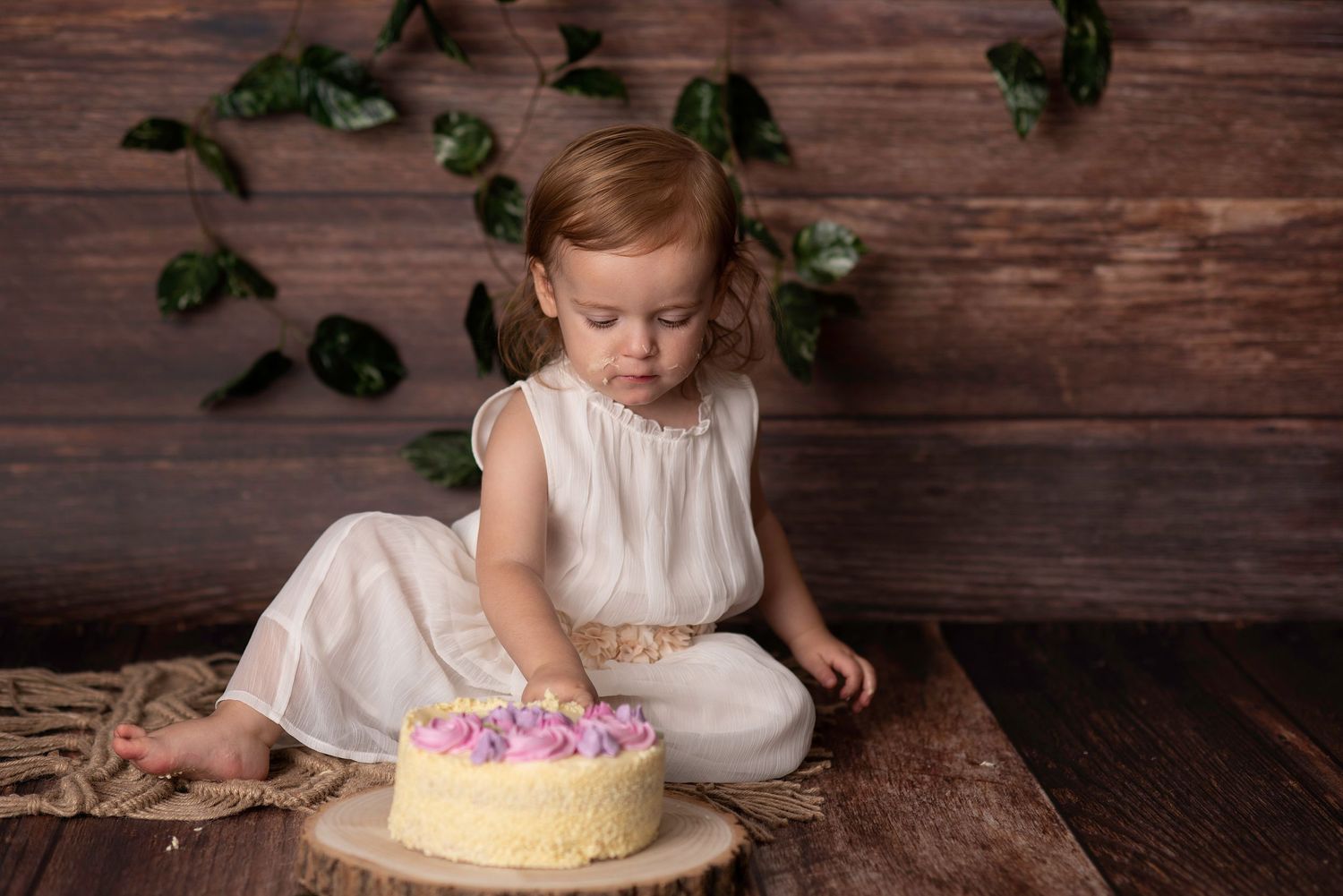 Small child in white dress explores a decorated cake on wooden floor with greenery backdrop.