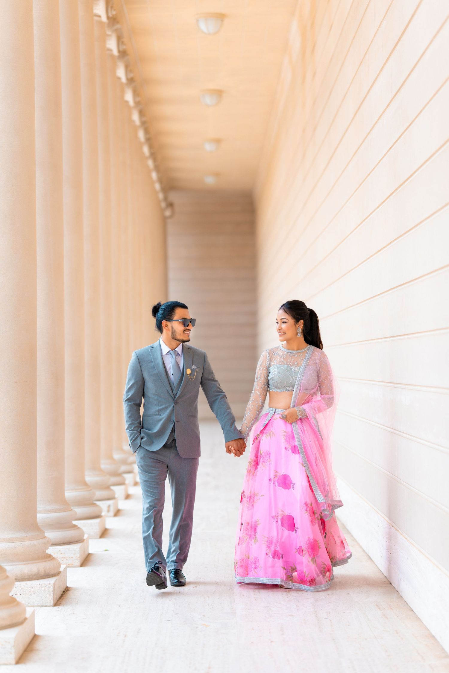 A couple strolls hand in hand through a grand colonnade wearing formal attire.