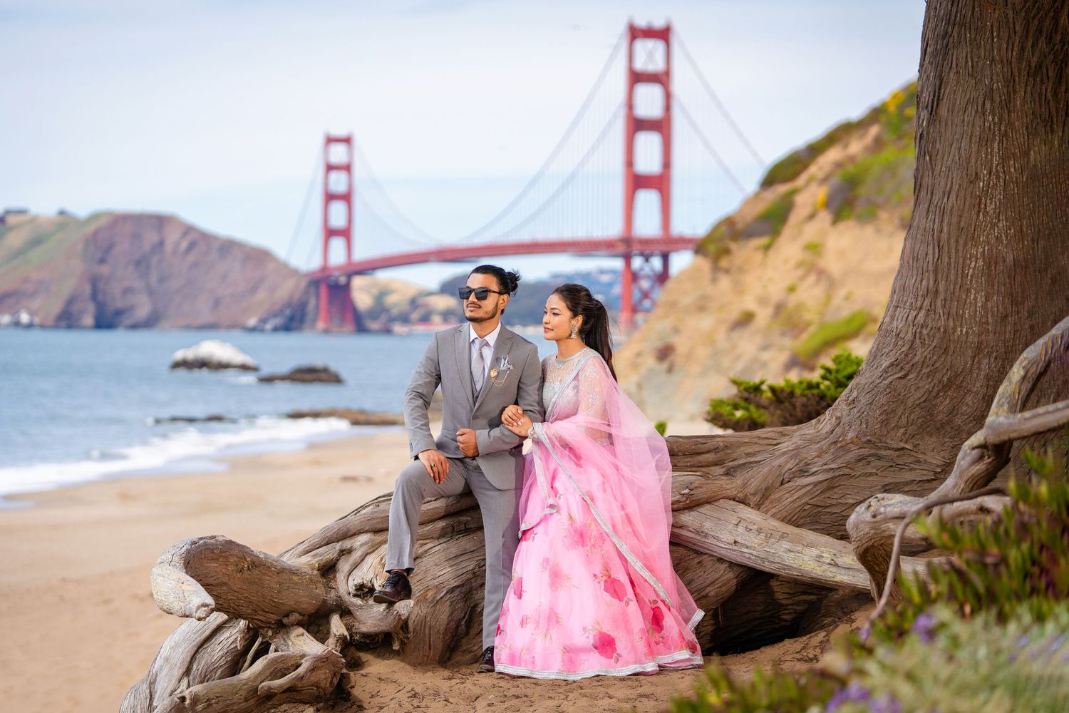 A couple in formal attire poses on driftwood with the Golden Gate Bridge as their backdrop.