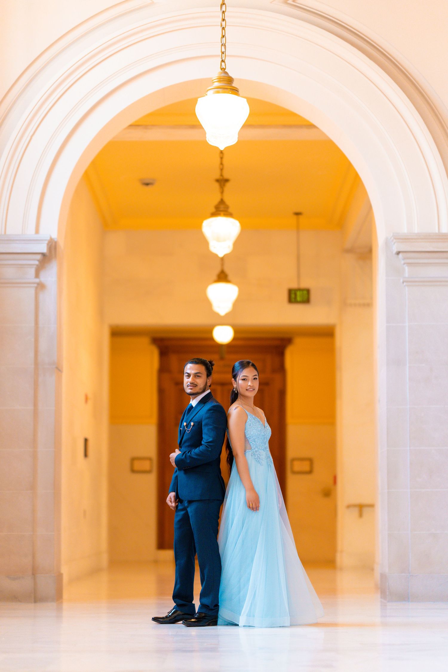 A formal couple poses in a hallway with pendant lights and arched architecture.