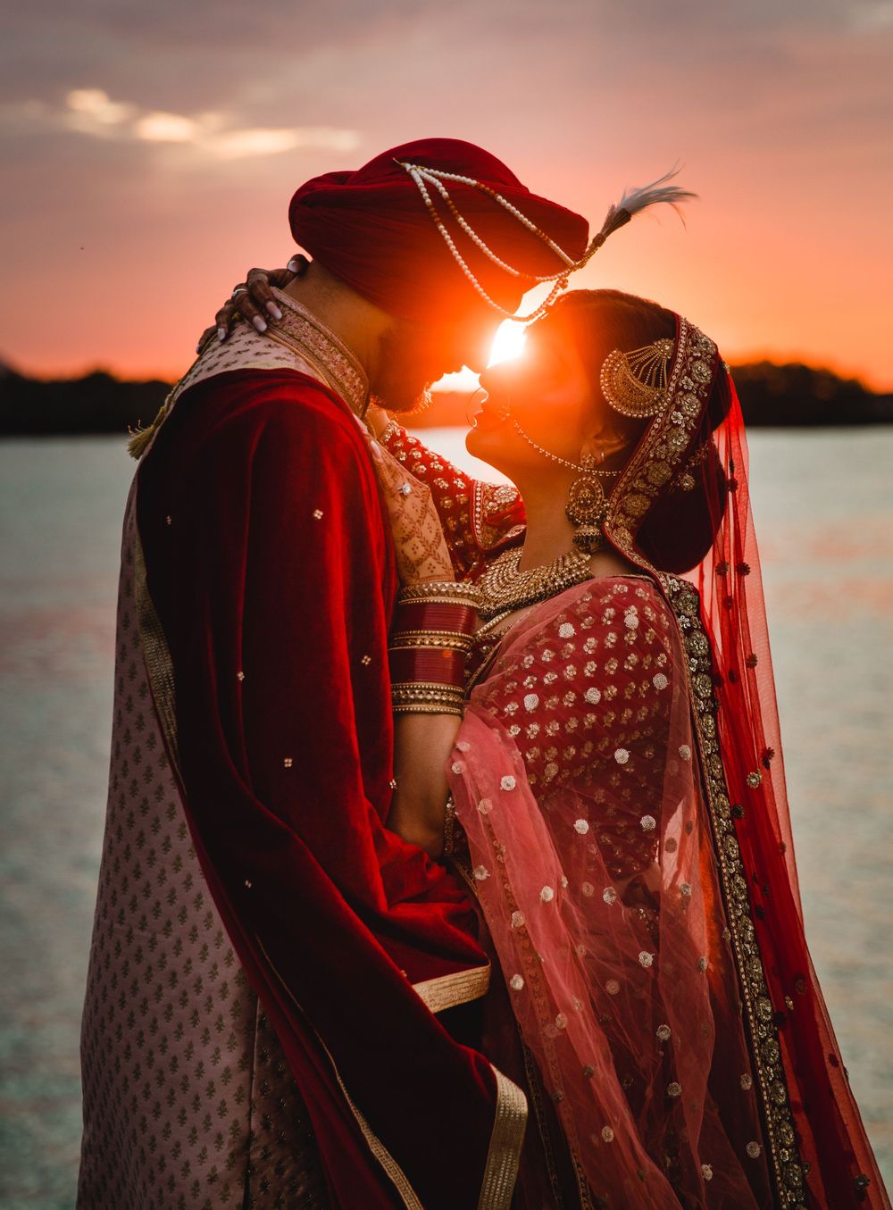 Silhouetted Indian wedding couple in traditional attire against a pink sunset over water.