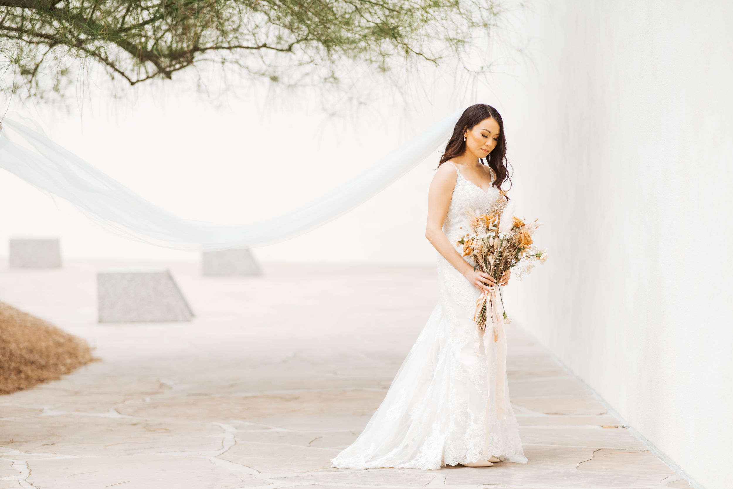 Bride in elegant white dress holds dried flower bouquet against white wall.