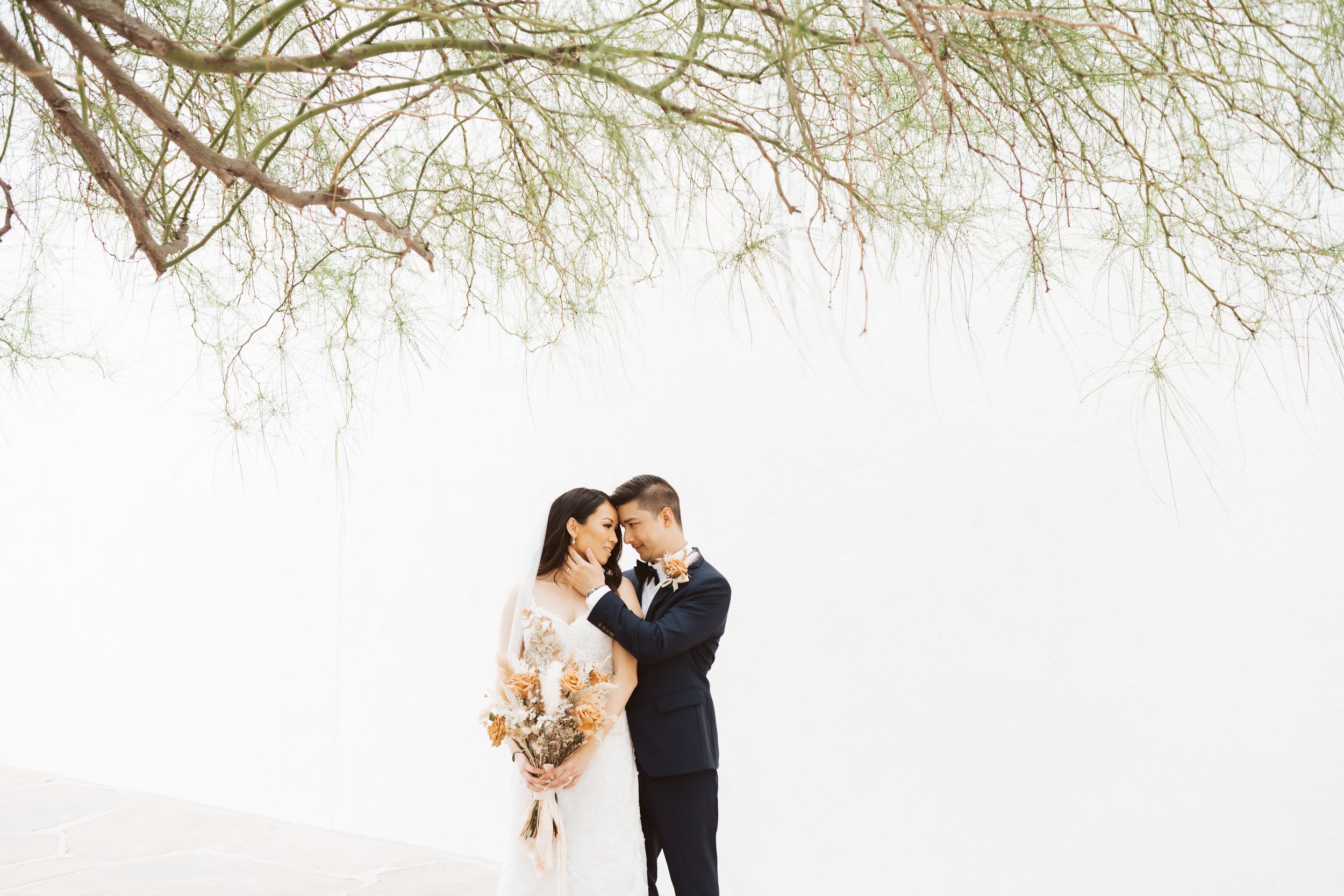Intimate black and white portrait of couple with dried flower bouquet against white wall.