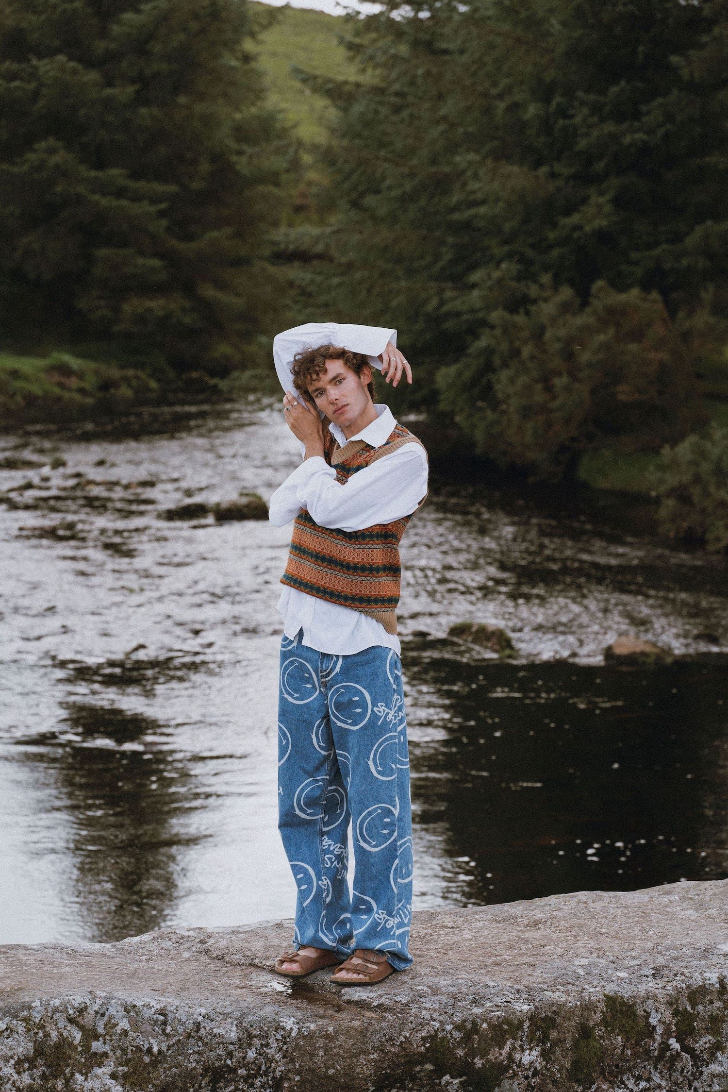 Someone poses artistically by a river wearing patterned blue jeans and a brown knit vest over white shirt.