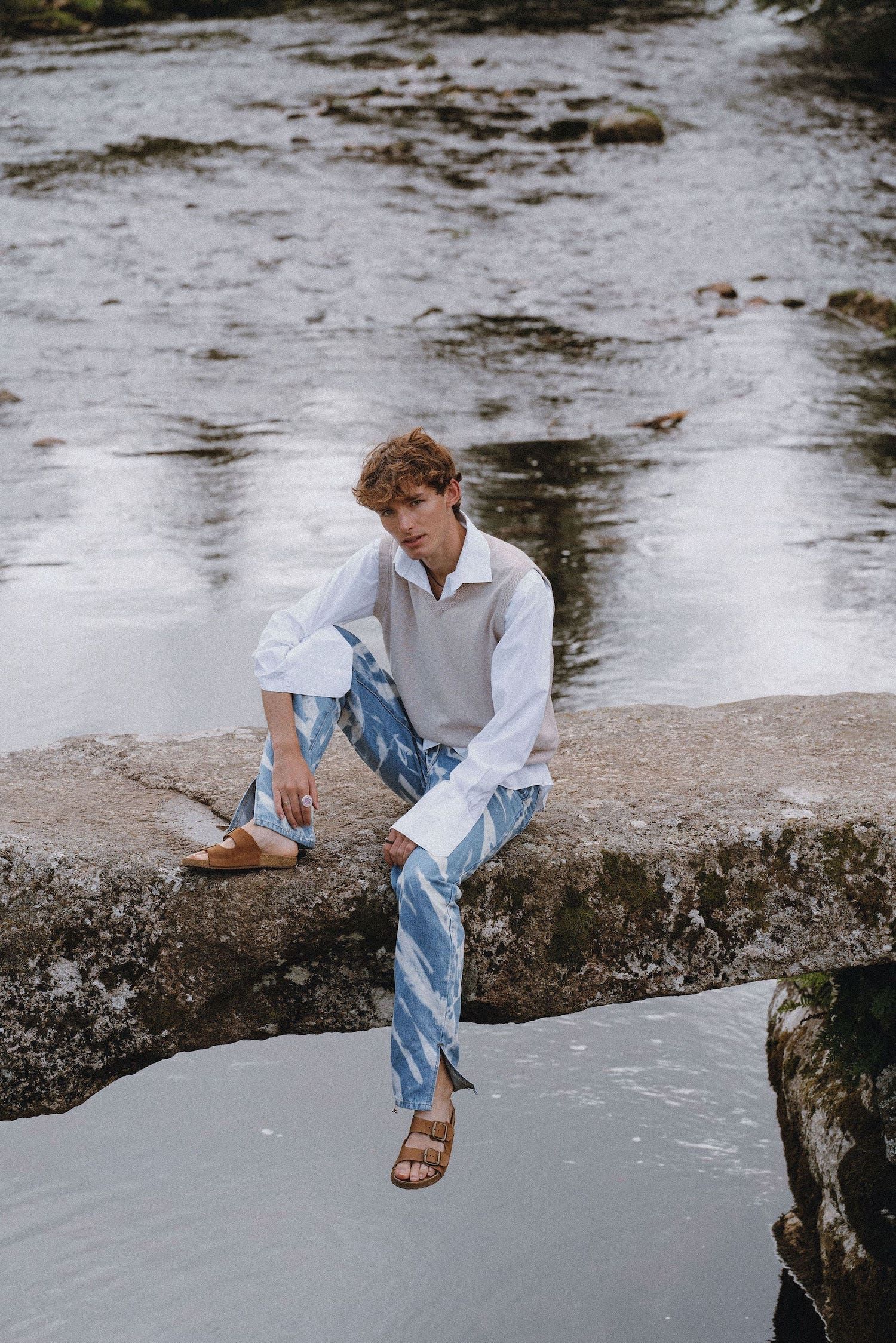 A casual outdoor portrait by a river showing someone in relaxed denim and sandals sitting on rocks.