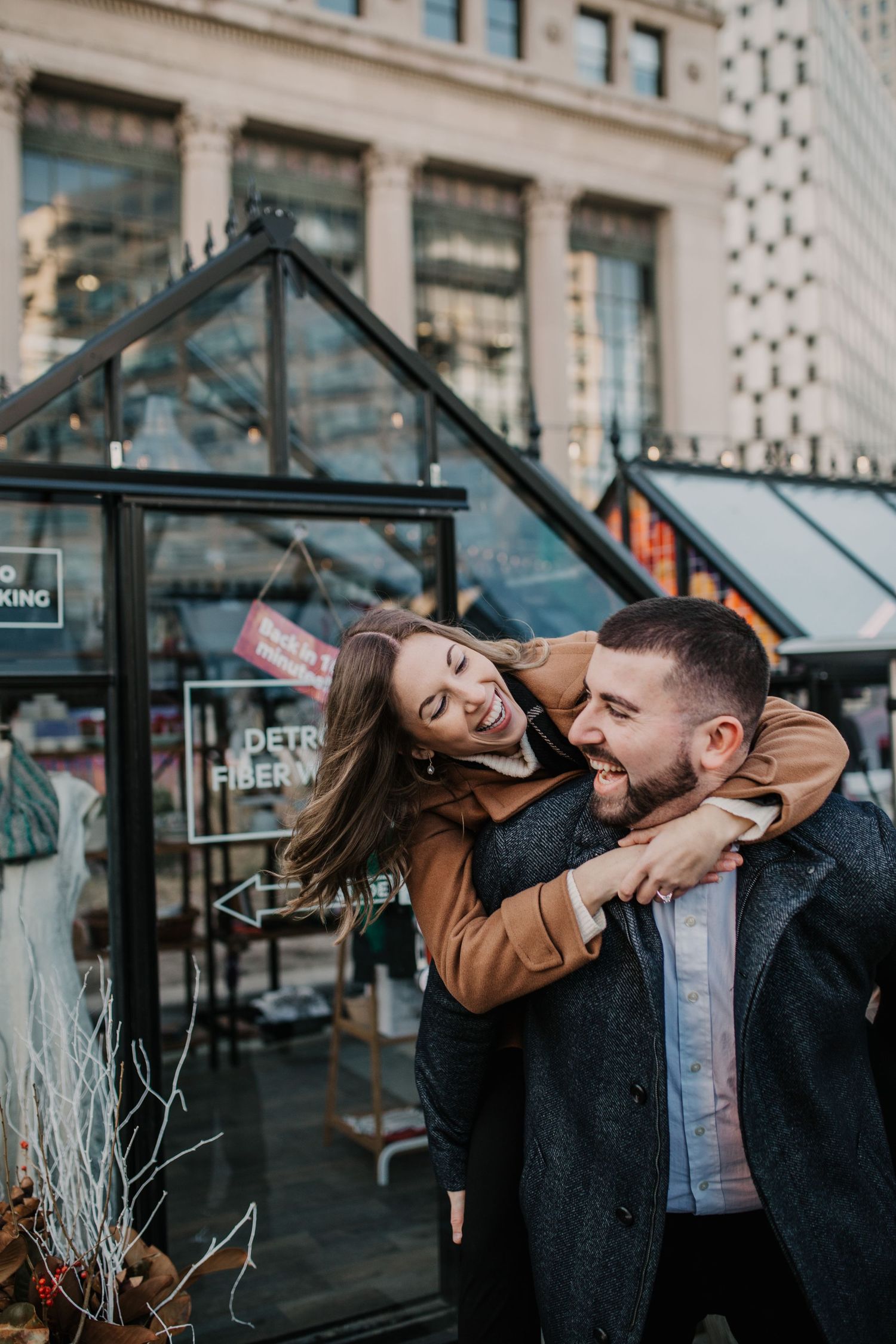 A playful couple laughs together outside a glass-fronted urban building.