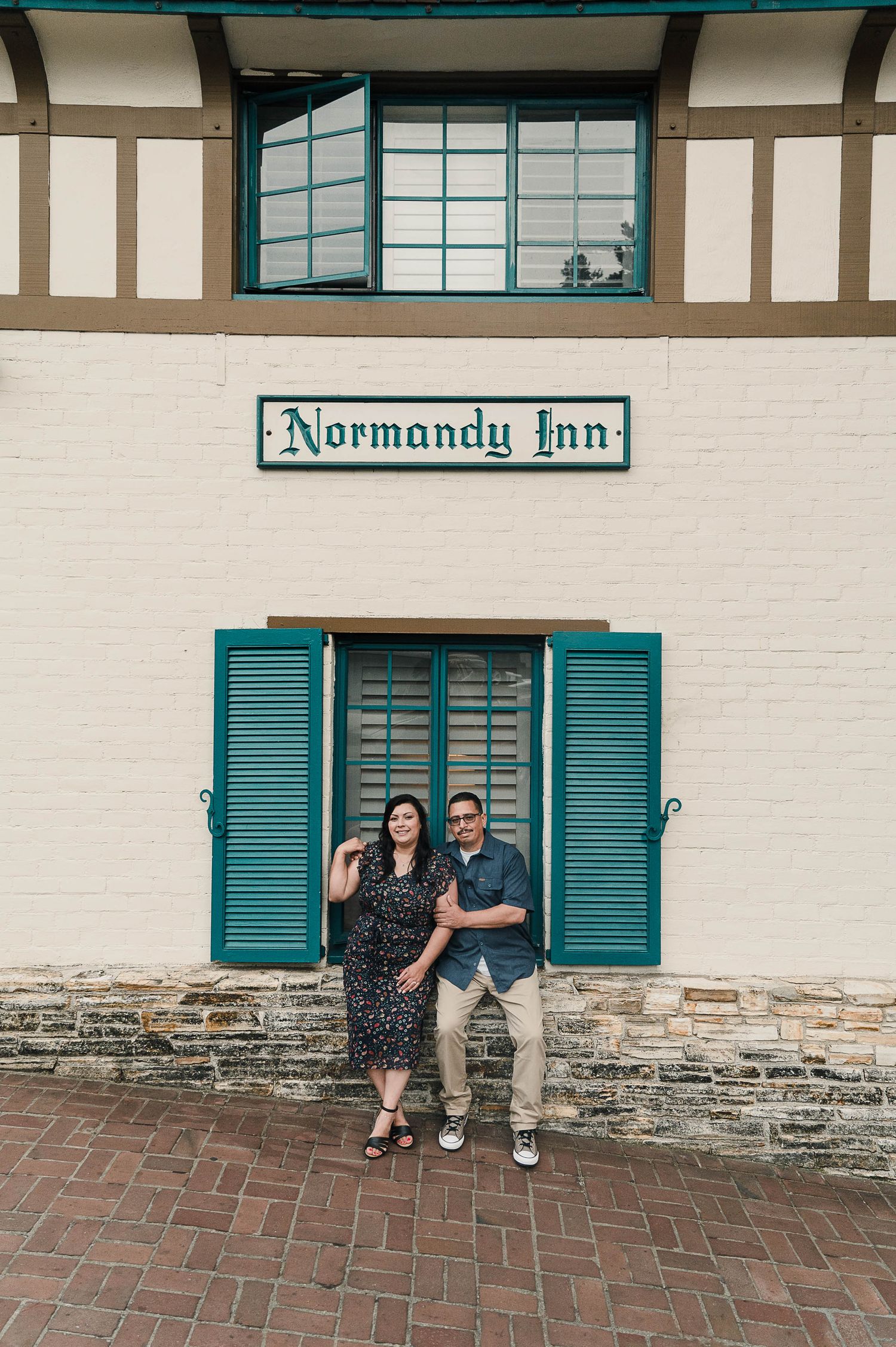 A couple poses in front of the Normandy Inn sign and teal shutters on a brick walkway.