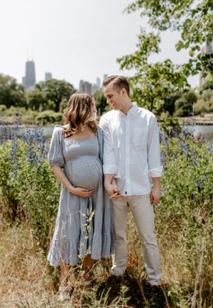 Expecting couple in casual attire standing in field of wildflowers with city backdrop.