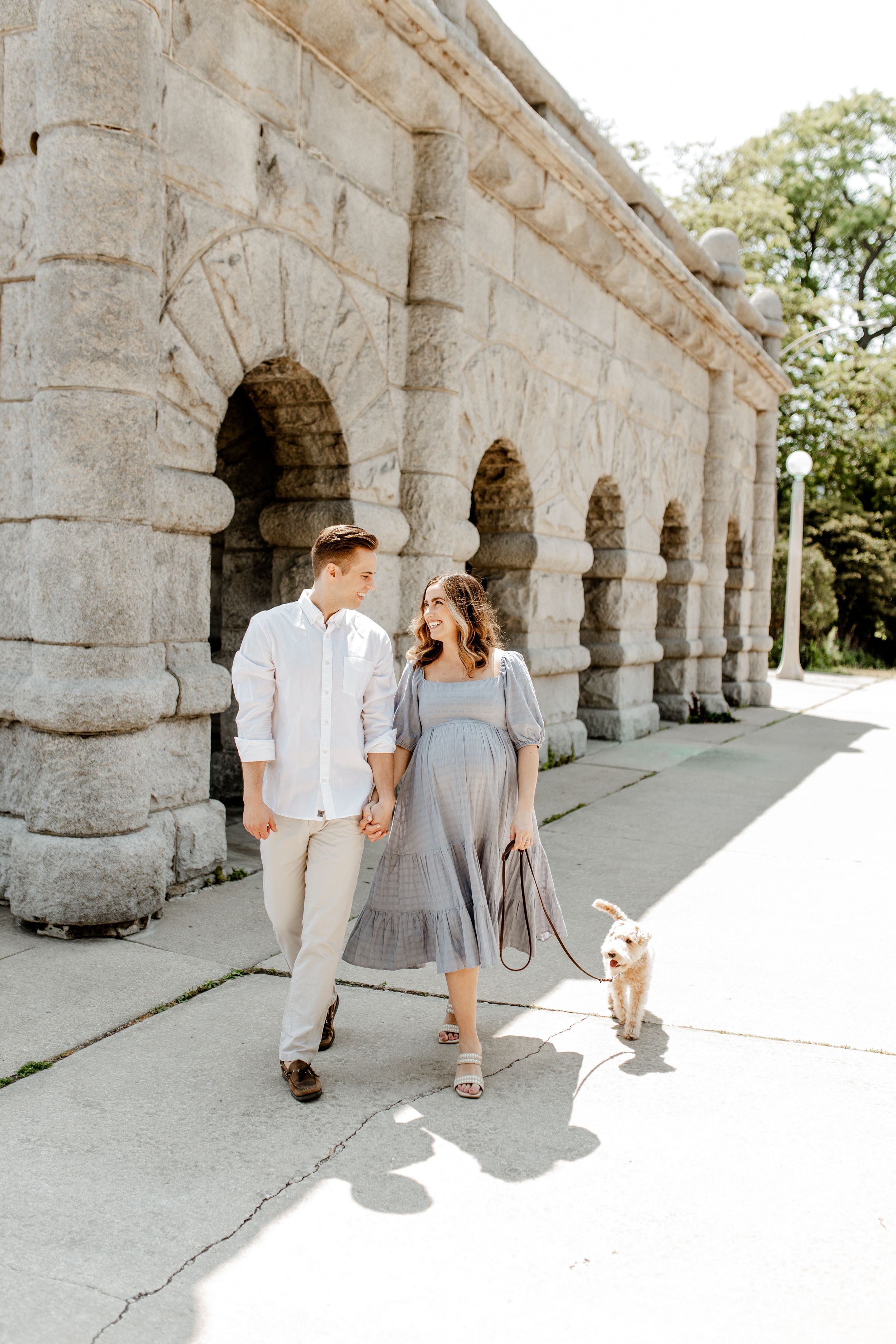 Couple walking with small dog along historic stone colonnade pathway.