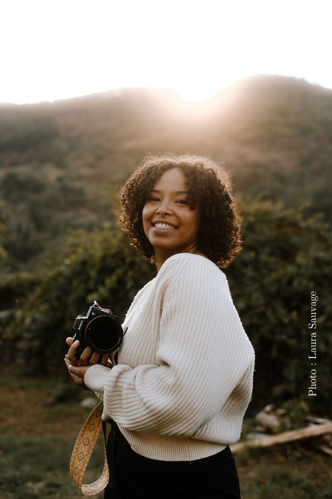 A photographer with curly hair holds a camera while smiling in warm sunset lighting.
