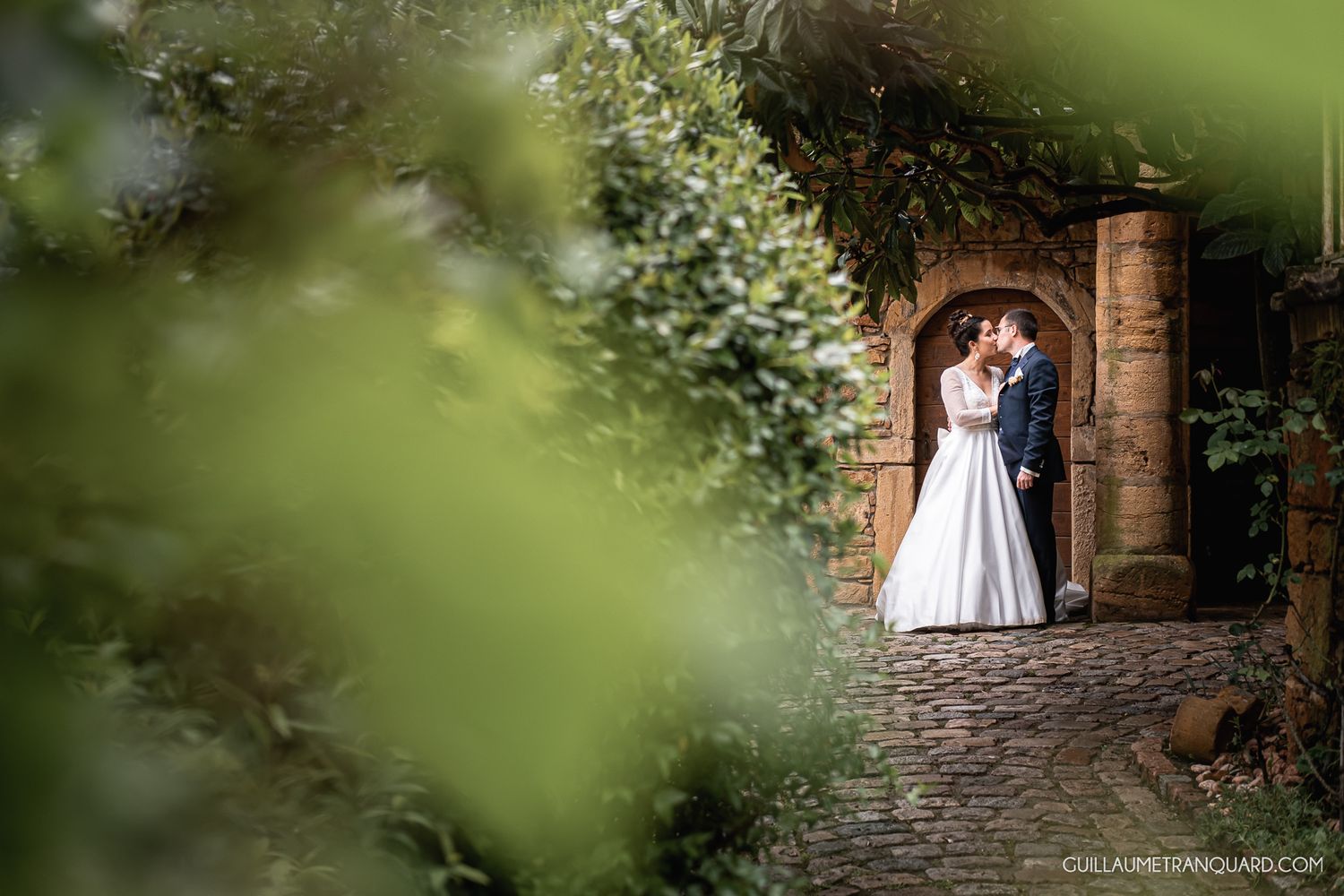 A white dress glimpsed through green foliage against stone architecture.
