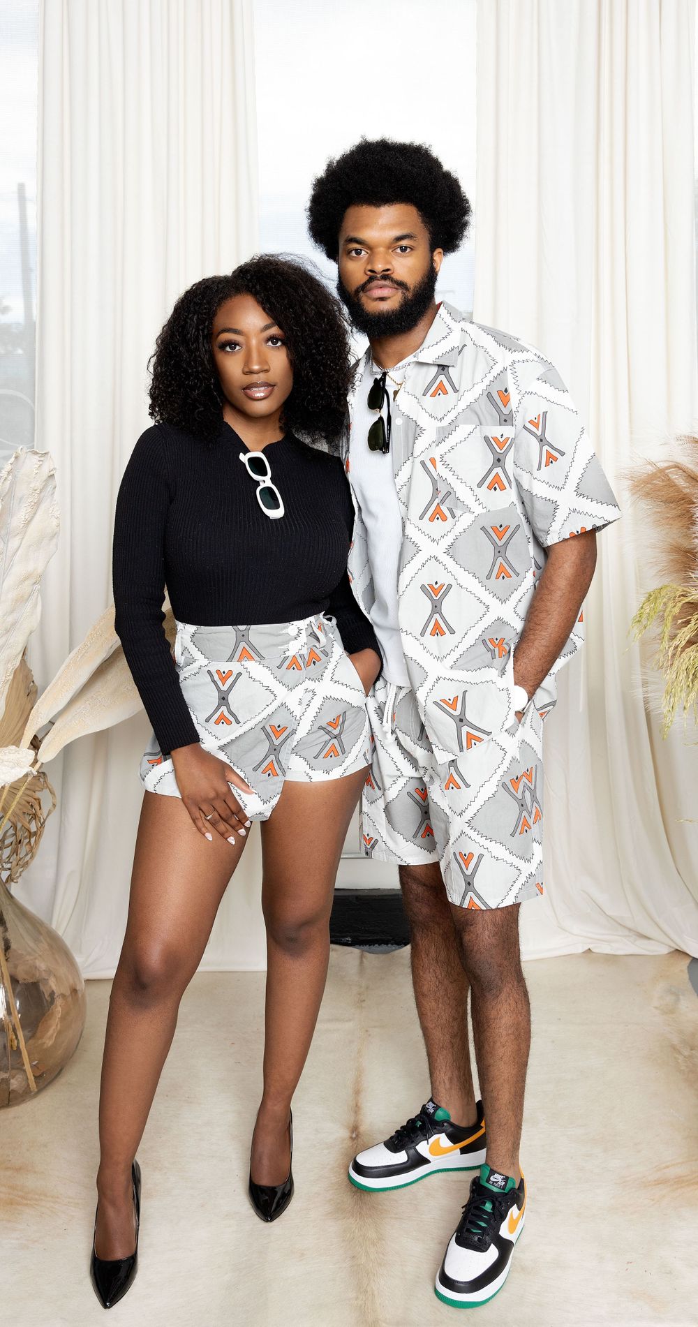 Couple wearing matching geometric patterned outfits poses together against white curtain backdrop.