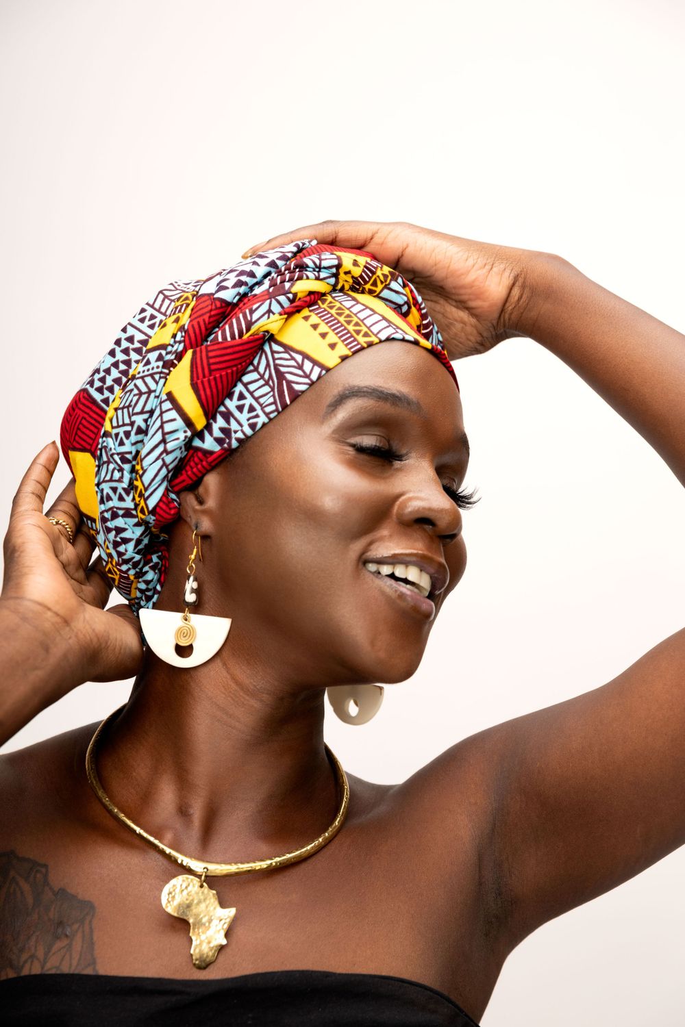 Person wearing colorful African print headwrap and gold necklace poses gracefully against white backdrop.