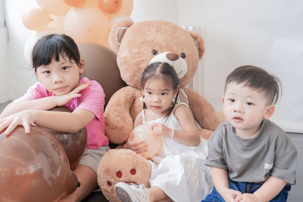 Children sit together with large teddy bears and peach-colored balloons in room.
