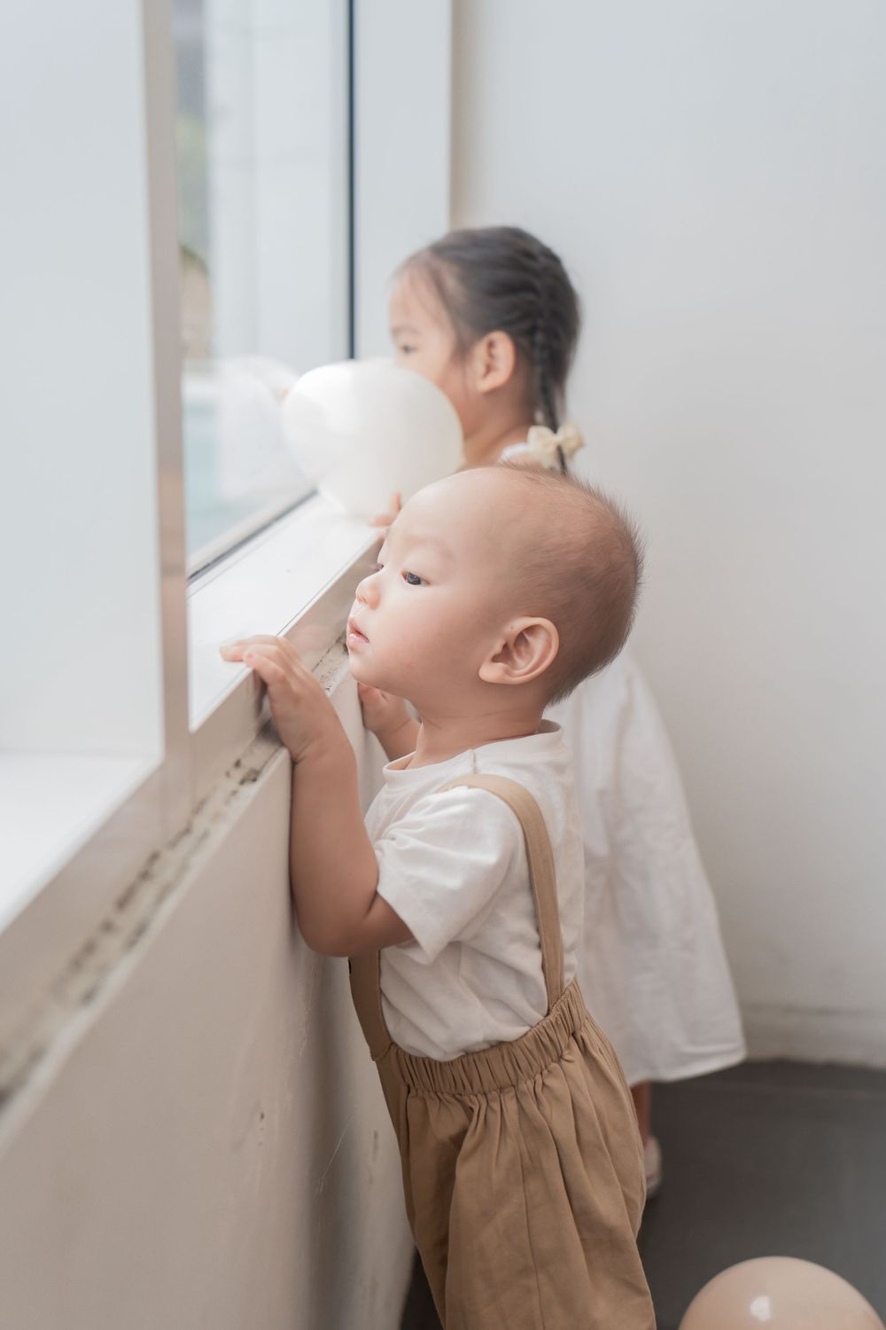 Two children peek out a bright window together in matching outfits.
