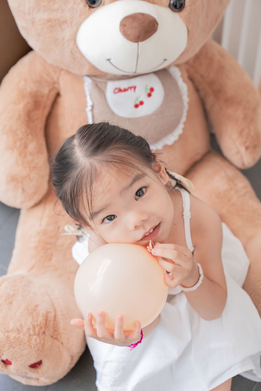 Child holds balloon while sitting in front of large teddy bear.