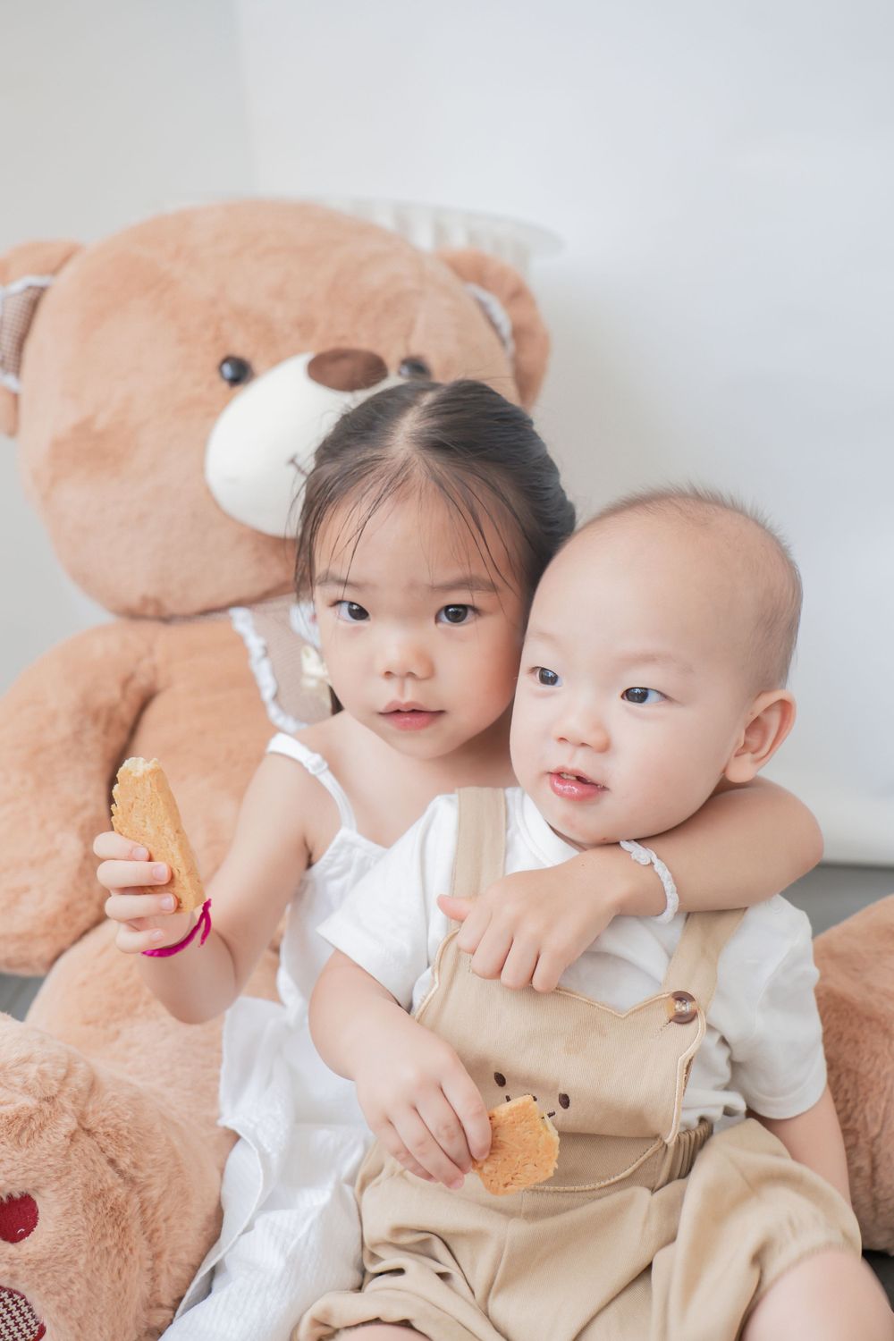 Two children pose with large teddy bear and snacks in bright room.