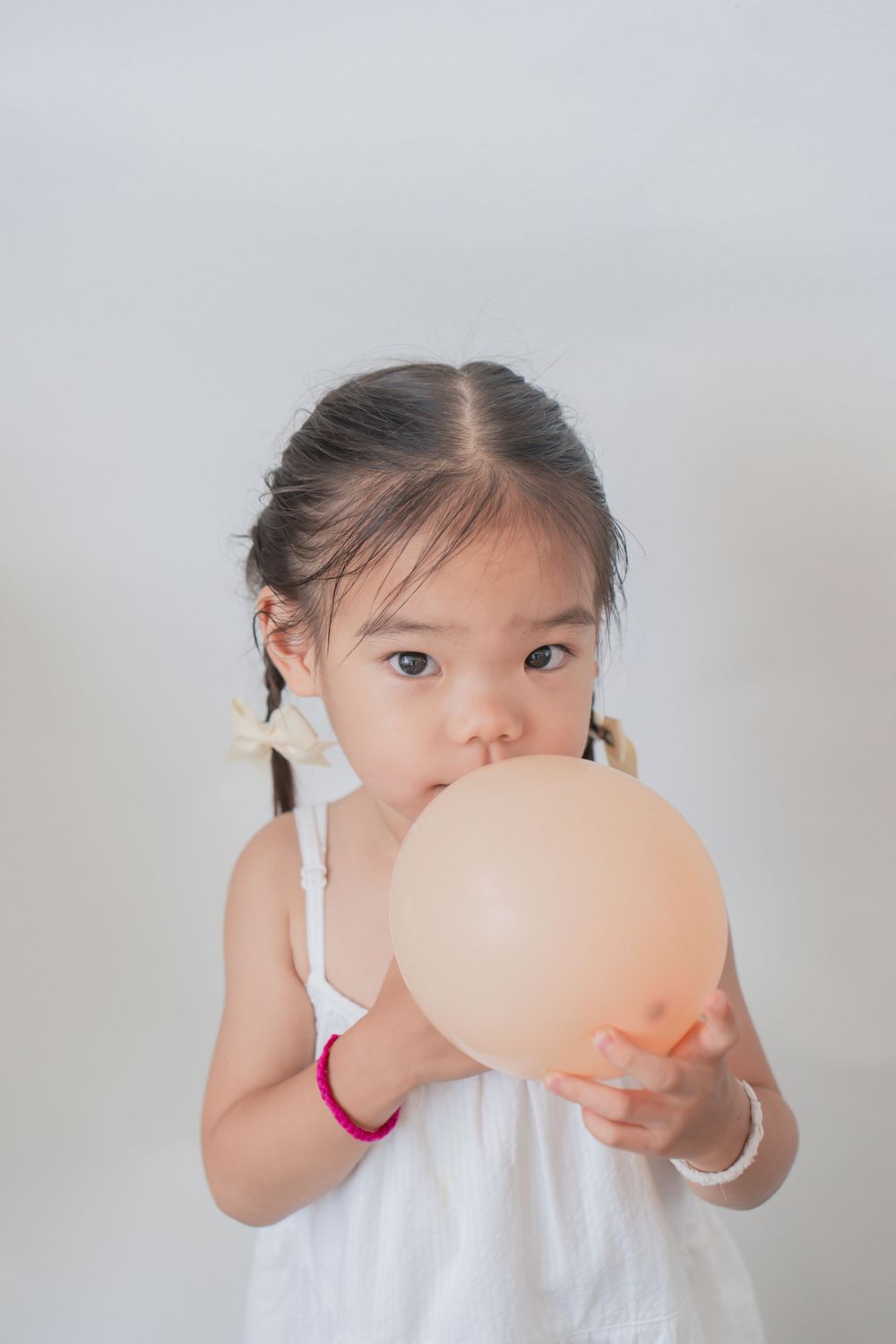 Child in white dress blows up peach-colored balloon.