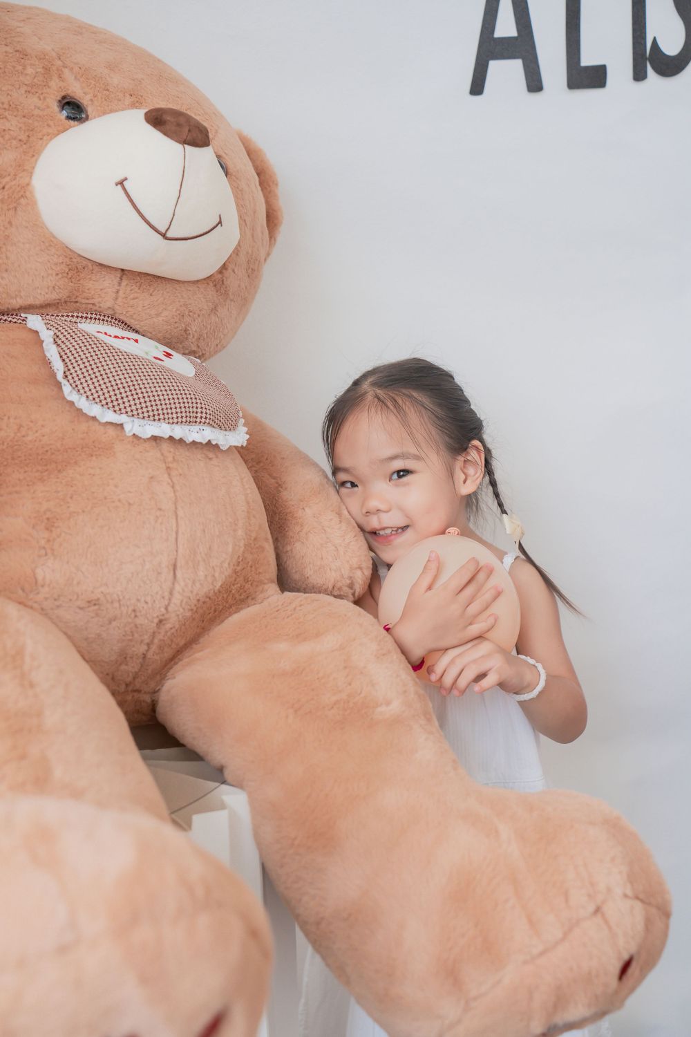 Child hugs large brown teddy bear against white wall with text.