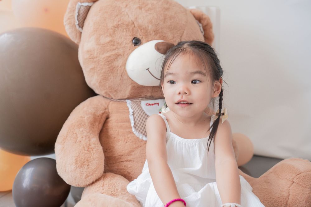 Child sits beside large brown teddy bear with white balloons in room.