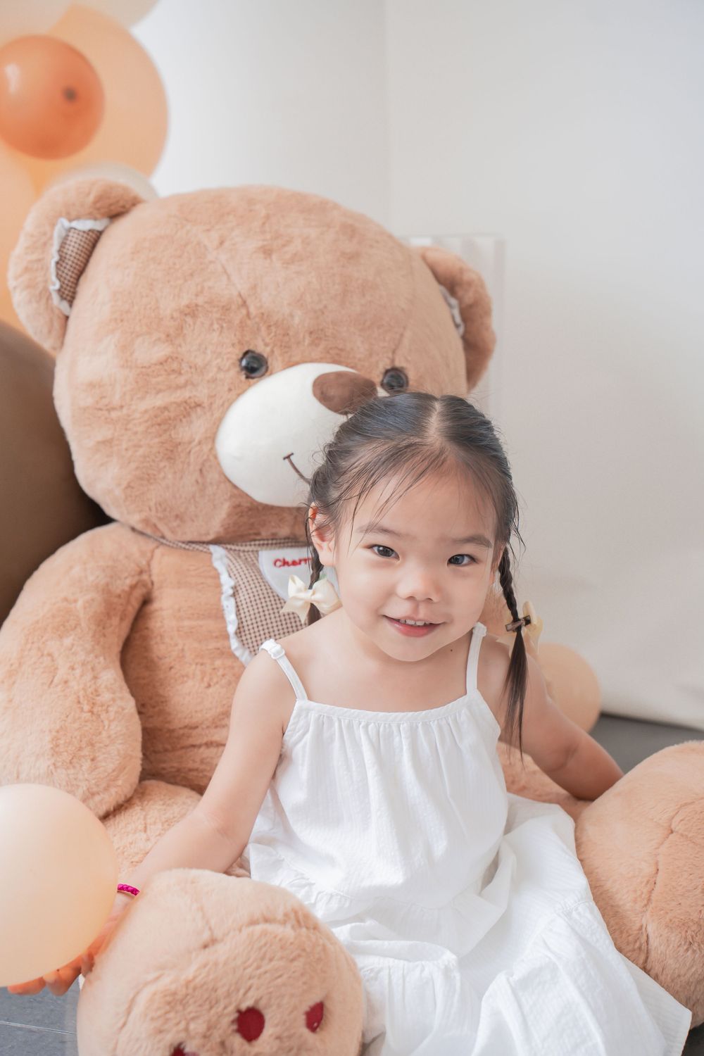 Child in white dress poses with oversized teddy bear against white wall.