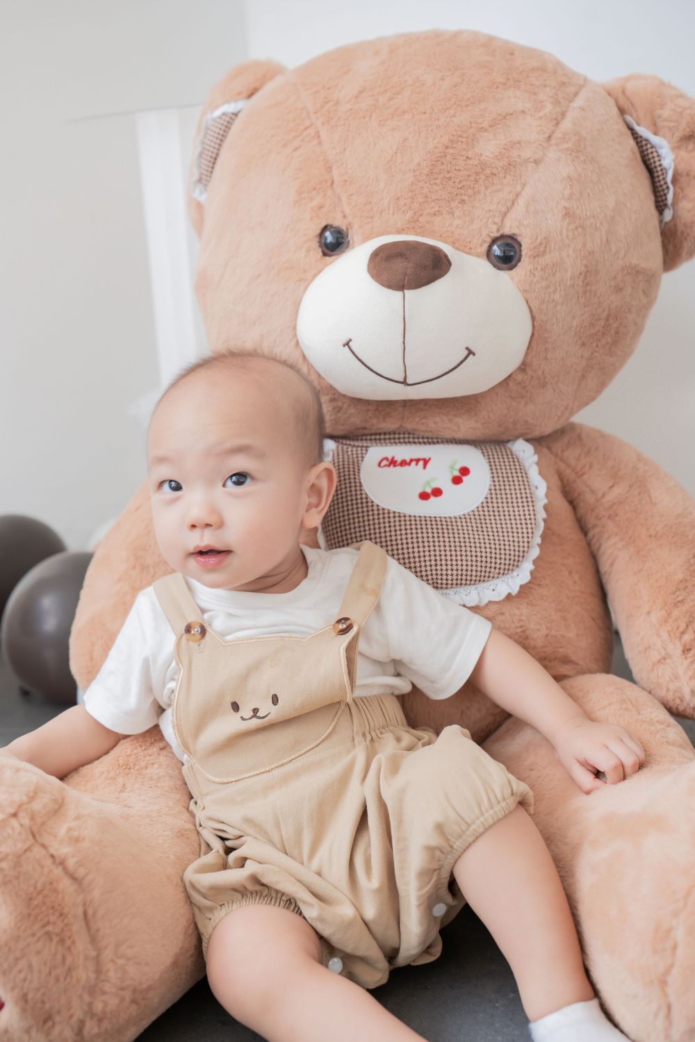 Baby in light colored overalls sitting with large plush teddy bear.