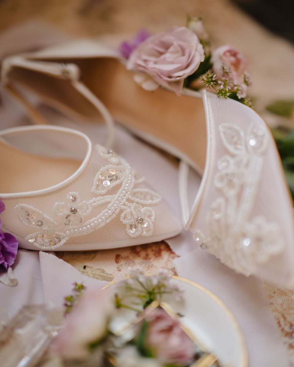 Ornate white bridal shoes decorated with beading and lace, paired with delicate pink and purple flowers.