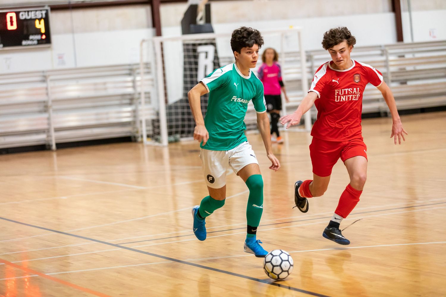 Youth futsal match between teams in red and green uniforms on indoor court.
