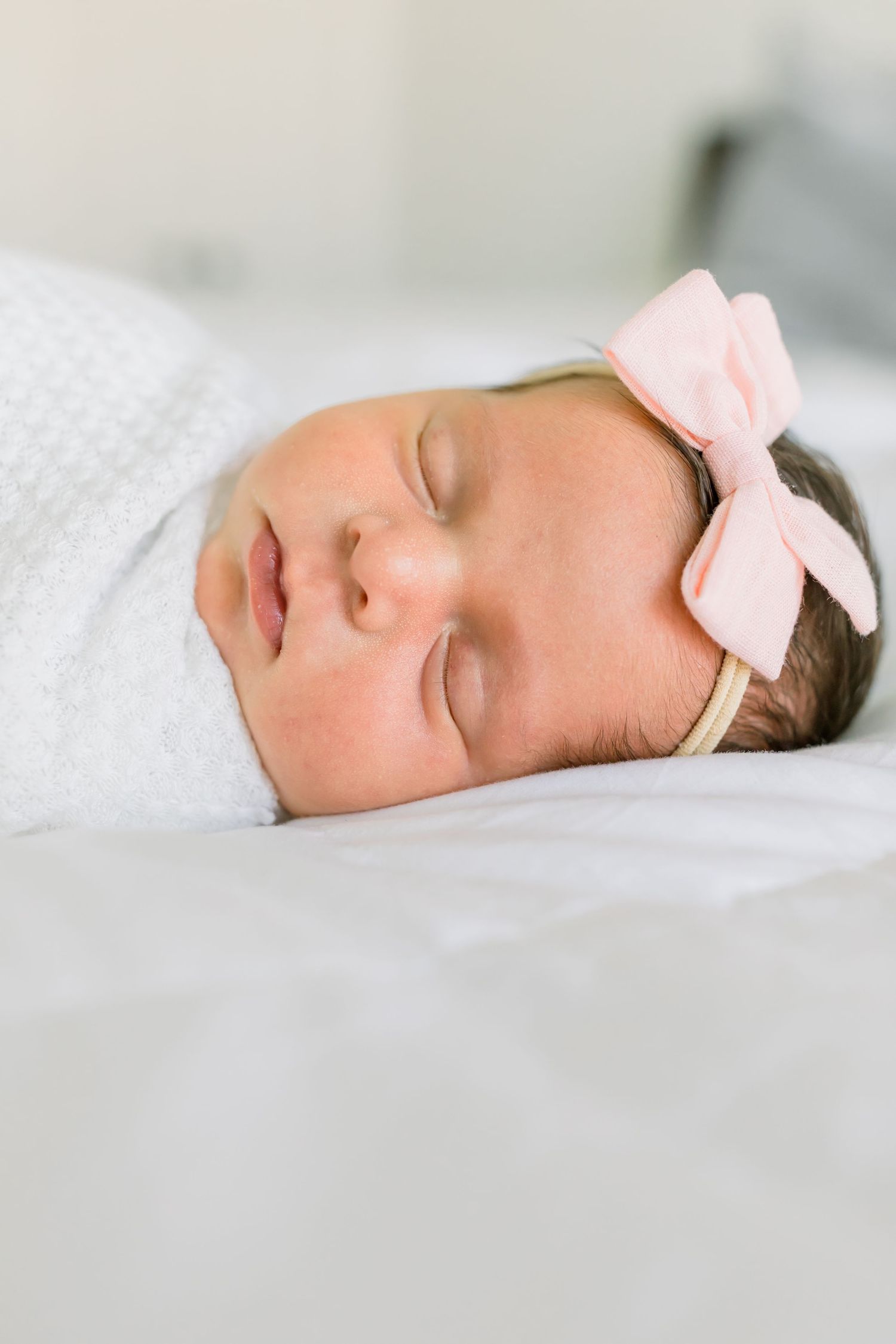 A sleeping baby wearing a pink bow headband swaddled in white.
