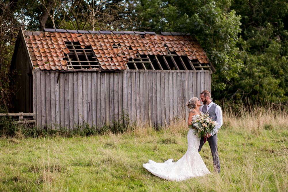 A wedding couple poses in front of a rustic weathered barn with a terracotta tile roof in a grassy field.